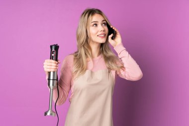Mor arka planda izole edilmiş el blenderi kullanan genç Rus kız elinde kahve ve cep telefonu var.