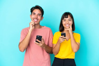 Mavi arka planda cep telefonu tutan melez bir çift tatlı bir ifadeyle gülümsüyor.