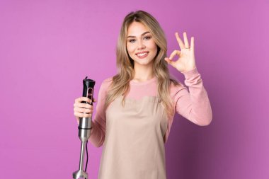 Mor arka planda izole edilmiş el blenderi kullanan genç Rus kız parmaklarıyla işaret veriyor.