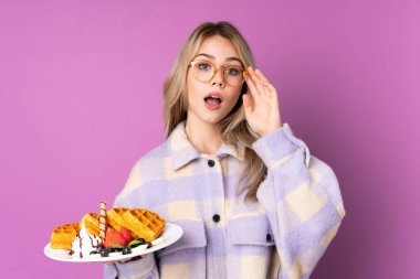 Mor arka planda gözlüklü ve şaşırmış bir şekilde waffle tutan genç Rus kız.