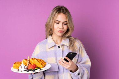 Elinde waffle tutan genç Rus kız mor arka planda izole edilmiş düşünüyor ve bir mesaj gönderiyor.