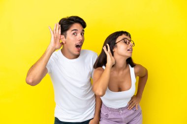 Genç melez çift sarı arka planda izole edilmiş bir şekilde ellerini kulağına koyup bir şeyler dinliyorlar.