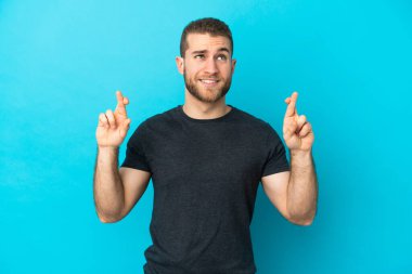 Mavi arka planda izole edilmiş genç, yakışıklı beyaz bir adam parmaklarını çapraz yapıp en iyisini diliyor.