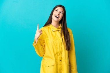 İzole edilmiş mavi arka planda yağmura dayanıklı bir ceket giyen Brezilyalı genç kız yukarı bakıyor ve şaşırıyor.