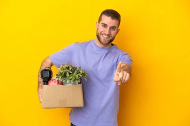 Genç Kafkasyalı, sarı arka planda izole edilmiş şeylerle dolu bir kutuyu kaldırırken ve parmağını kaldırırken...