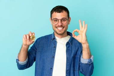 İzole edilmiş mavi arkaplan üzerinde Bitcoin tutan Brezilyalı bir adam tamam işaretini parmaklarıyla gösteriyor