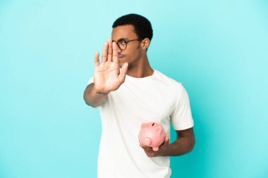 Afro-Amerikalı yakışıklı bir adam mavi arka plan üzerinde bir kumbara tutuyor dur hareketi yapıyor ve hayal kırıklığına uğruyor.