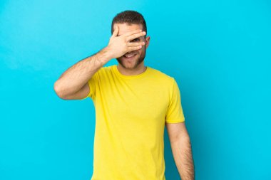 Genç yakışıklı beyaz adam mavi arka planda izole edilmiş gözleri elleriyle kaplıyor ve gülümsüyor.
