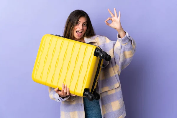 度假时 年轻的高加索女人带着旅行箱被隔离在紫色的背景下 并制作了Ok标志 — 图库照片