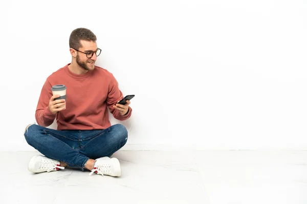 年轻英俊的男子坐在地板上 手里拿着咖啡要带走 手里拿着手机 — 图库照片