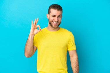 Mavi arka planda izole edilmiş yakışıklı beyaz adam parmaklarıyla işaret veriyor.