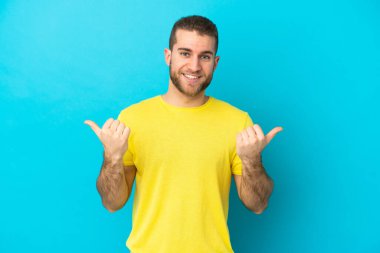Mavi arka planda soyutlanmış yakışıklı beyaz bir adam. Baş parmakları havada ve gülümsüyor.