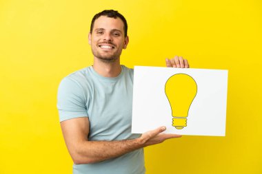 İzole edilmiş mor arka plan üzerinde Brezilyalı bir adam elinde mutlu ifadeli bir ampul simgesi olan bir levha tutuyor.