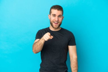 Mavi arka planda izole edilmiş yakışıklı beyaz adam şaşırmış ve ön tarafı işaret ediyor.