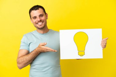 İzole edilmiş mor arka plan üzerinde Brezilyalı bir adam elinde ampul simgesi olan bir levha tutuyor ve ona doğrultuyor.