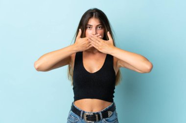 Mavi arka planda izole edilmiş genç beyaz kadın elleriyle ağzını kapatıyor.