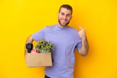 Genç Kafkasyalı, sarı arka planda izole edilmiş eşyalarla dolu bir kutuyu kaldırırken hareket ediyor.