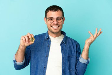 Brezilya 'lı bir adam Bitcoin' i izole edilmiş mavi arka planda tutuyor. Davet etmek için ellerini yana uzatıyor.