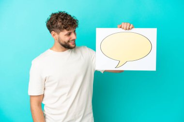 Genç beyaz adam mavi arka planda izole edilmiş. Elinde konuşma balonu ikonu olan bir levha var.