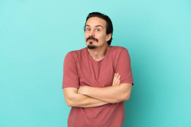 Kafkasyalı genç adam mavi arka planda izole edilmiş. Omuzlarını kaldırırken şüpheyle hareket ediyor.