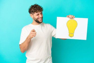 Genç beyaz adam mavi arka planda izole edilmiş. Elinde ampul simgesi olan bir levha var ve ön tarafı işaret ediyor.