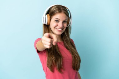 Genç beyaz kadın mavi arka planda izole edilmiş müzik dinliyor ve ön tarafı işaret ediyor.