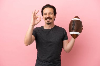 Pembe arka planda ragbi oynayan beyaz bir adam parmaklarıyla işaret veriyor.