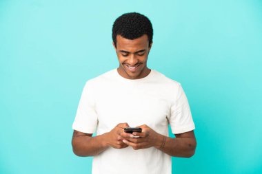 İzole edilmiş mavi arka planda Afro-Amerikalı yakışıklı bir adam cep telefonuyla mesaj gönderiyor.