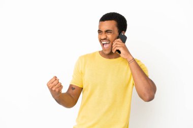 Afro-Amerikalı yakışıklı adam, izole edilmiş beyaz arka plan üzerinde cep telefonu kullanıyor ve zaferi kutluyor.