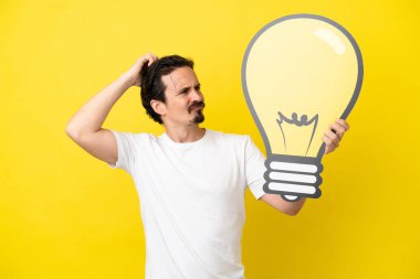 Sarı arka planda izole edilmiş genç beyaz bir adam elinde bir ampul simgesi ve şüpheleri var.