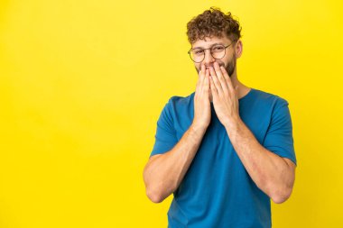 Sarı arka planda izole edilmiş yakışıklı beyaz adam mutlu ve gülümseyen elleriyle ağzını kapatıyor.