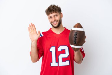 Beyaz arka planda ragbi oynayan beyaz tenli genç adam mutlu bir ifadeyle selamlıyor.