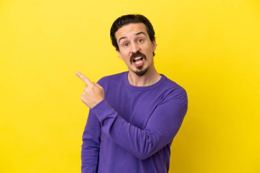 Sarı arka planda izole edilmiş genç beyaz adam şaşırmış ve işaret ediyor.