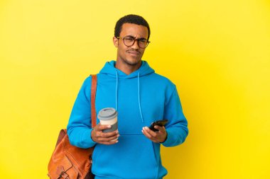 Afrika kökenli Amerikalı bir öğrenci sarı arka planda, elinde kahve ve bir şeyler düşünürken cep telefonu tutuyor.