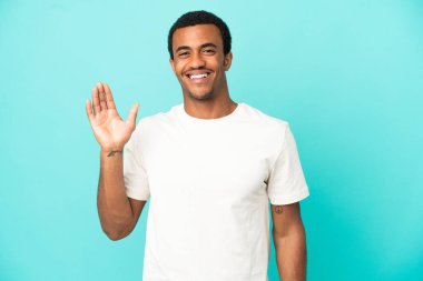 İzole edilmiş mavi arka planda Afro-Amerikalı yakışıklı adam mutlu bir ifadeyle selamlıyor.