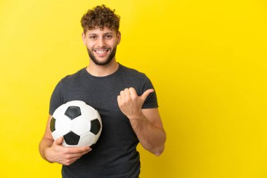 Yakışıklı genç futbolcu sarı arka planda izole edilmiş bir şekilde bir ürün sunmak için kenarı işaret ediyor.