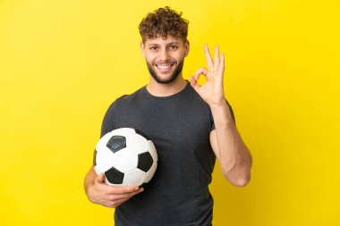 Sarı arka planda izole edilmiş yakışıklı genç futbolcu parmaklarıyla tamam işareti gösteriyor