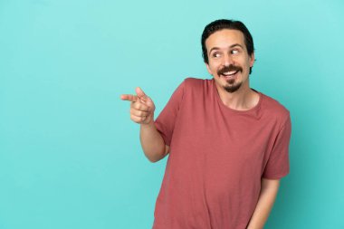 Mavi arka planda izole edilmiş genç beyaz bir adam parmağını yana doğru uzattı ve bir ürün sundu.