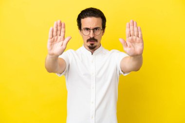 Sarı arka planda izole edilmiş genç beyaz adam dur hareketi yapıyor ve hayal kırıklığına uğruyor.