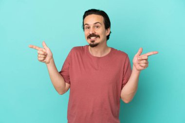 Mavi arka planda izole edilmiş genç beyaz adam parmak uçlarını işaret ediyor ve mutlu.