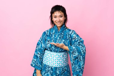 İzole edilmiş mavi arka planda kimono giyen genç bir kadın gülümseyerek bir fikir sunuyor.