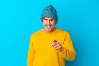 Yakışıklı sarışın adam mavi arka planda izole edilmiş, şaşırmış ve ön tarafı işaret ediyor.