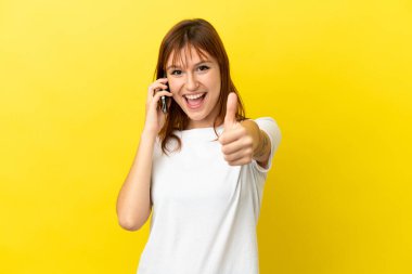 Kızıl saçlı kız sarı arka planda izole edilmiş. Başparmaklarını kaldırırken cep telefonuyla sohbet ediyor.