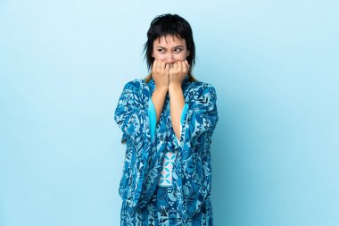 İzole edilmiş mavi arka planda kimono giyen genç bir kadın gergin ve eli ağıza vermeye korkuyor.