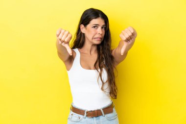 Sarı arka planda izole edilmiş genç beyaz kadın baş parmağını iki eliyle aşağı gösteriyor.