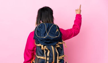 İzole edilmiş pembe arka planı olan büyük sırt çantalı genç dağcı kız işaret parmağıyla işaret ediyor.