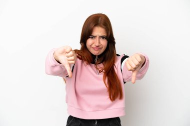 Beyaz arka planda izole edilmiş genç kızıl saçlı kadın baş parmağını iki eliyle aşağı gösteriyor.