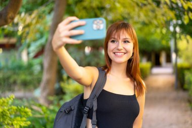 Genç, güzel, kızıl saçlı, açık havada selfie çeken bir kadın.