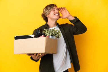 Sarışın adam, sarı arka planda izole edilmiş şeylerle dolu bir kutuyu kaldırırken çok fazla gülümsüyor.