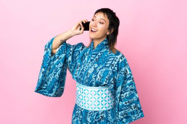 Kimono giyen genç bir kadın izole edilmiş mavi arka planda cep telefonuyla sohbet ediyor.